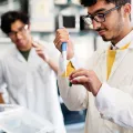 Student with medium skin tone and dark hair wearing black glasses and white lab coat learns how to use special laboratory equipment with professor watching in background