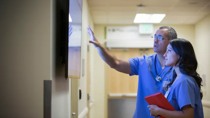 A medium skin toned man and a medium skin toned woman, both in blue scrubs, look at a wall-mounted screen. He is speaking and gesturing at the screen.