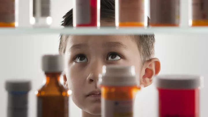 A young, medium skin-toned boy looks up at shelves of prescription medicine bottles