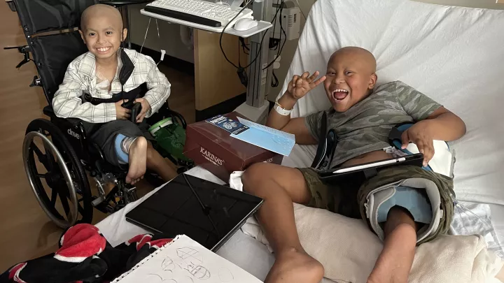 Two boys smile in a hospital room. One is in bed; one is in a wheelchair. Each boy has one shorter leg with a foot that points down.