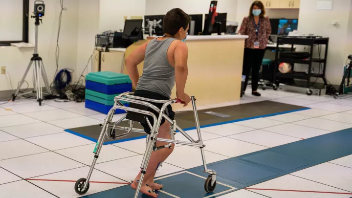 A boy with short dark hair walks with a walker across a blue-tiled path in a gait lab. He has sensors on his feet and legs.