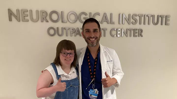 Medium shot of a light-skinned young adult woman in overalls posing with a light-skinned male doctor and giving the thumbs-up in a hospital front of a sign that reads “Neurological Institute Outpatient Center”