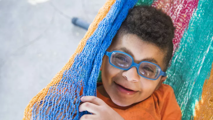 A medium-dark skin-toned boy wearing blue glasses smiles while laying in a colorful hammock