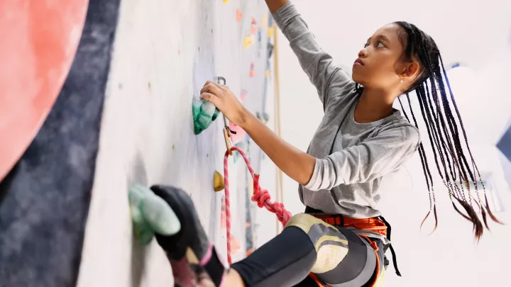 Teen girl with medium skin and long, thin black braids climbs a rock-climbing wall. One hand and one foot are on two green holds.