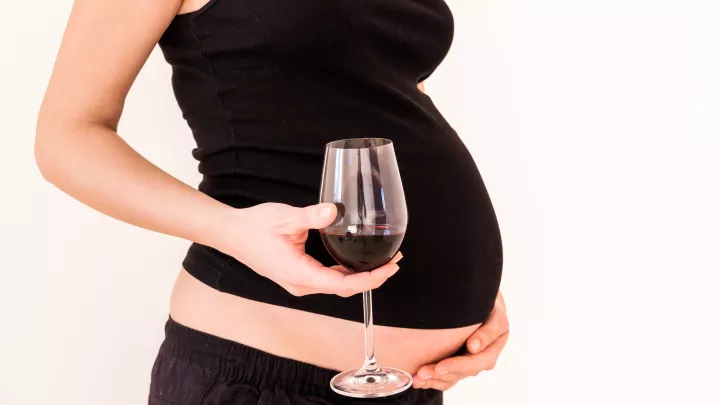 The torso of a light skin-toned pregnant woman, holding a glass of wine in her hand