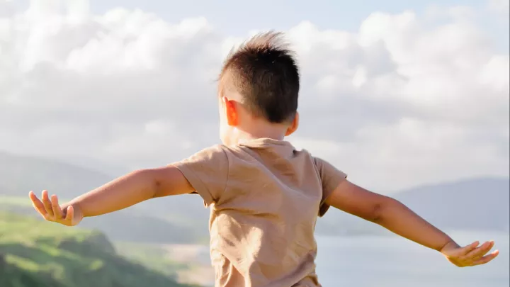 Photo taken from behind of a young boy with medium-light skin tone spreading his arms wide and standing on a hilltop with a view of green hills, the ocean, and white clouds in a blue sky in the background.
