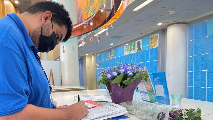 A medium skin-toned man in a blue polo shirt and surgical mask writes in a book