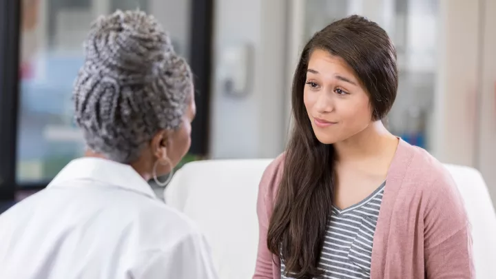 Young woman with medium skin and long dark hair sits on an exam table across from a female doctor whose back is turned. 