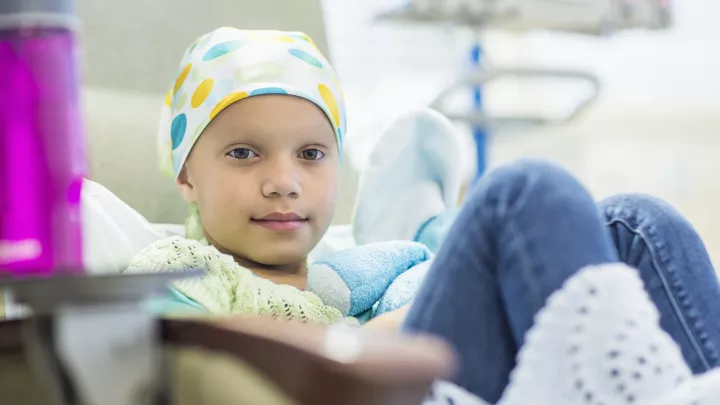 A child wearing a white headscarf with yellow and blue circles on it sits in a hospital bed, looking at the camera.