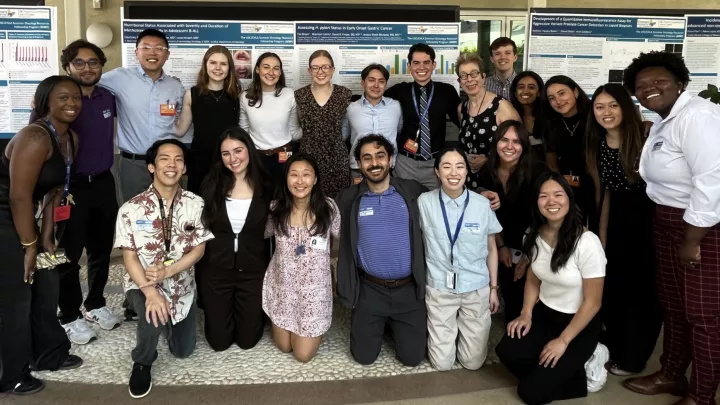 Group shot of adults in formal attire, with a variety of skin tones, posing in front of several scientific posters in an outdoor patio.