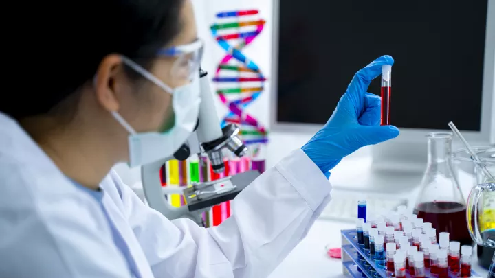 Scientist in white coat holds a small tube of blood with a blue-gloved hand. A colorful DNA strand model is in the background.