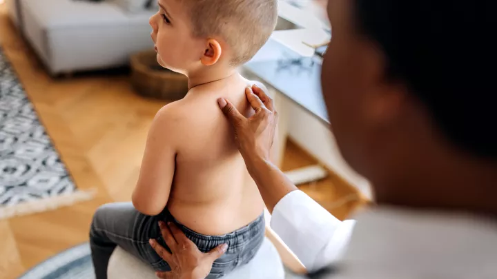 Rear view of a female doctor examining a boy’s back, with one hand at the top of the boy’s spine and one on his hip.