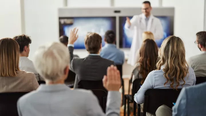 Man with light skin tone wearing lab coat and glasses instructs group of professionally dressed adult students in classroom setting