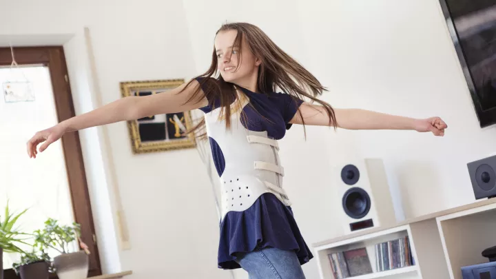 Light-skinned teen girl smiles and spins, arms outstretched, while wearing a white back brace over her shirt. 