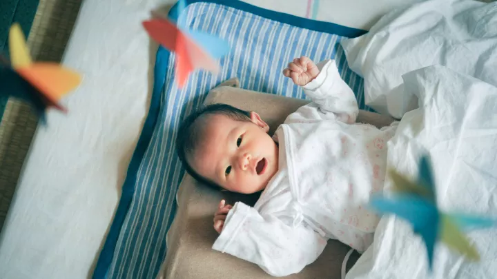 An infant with dark hair and eyes lies on his back on a blanket, looking up in wonder at a colorful mobile spinning above him. 