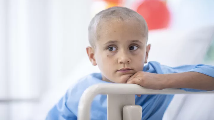 A young boy with a bald head, wearing a blue hospital gown, leans his head against the hospital bed railing. 