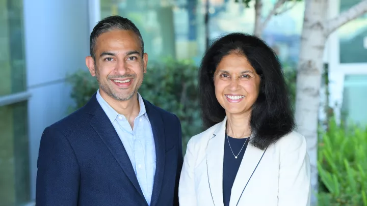 A man in a blue suit jacket and a woman wearing a light blazer stand together smiling. Both have dark hair and medium skin.