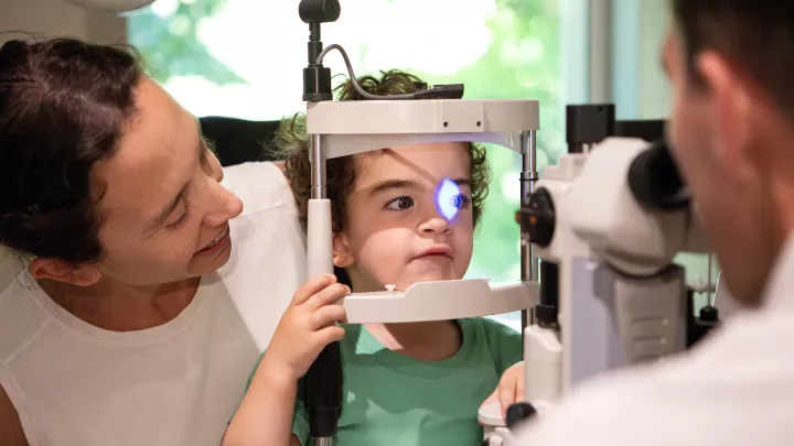 A light shines in a young child’s eyes as an unseen ophthalmologist checks her vision. The child is sitting on her mother’s lap.