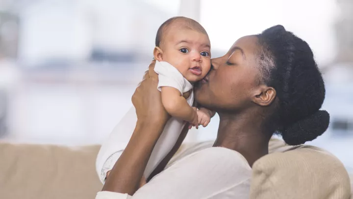 A dark-skinned mother holds her baby high and kisses her baby on the cheek. The baby is looking at the camera.