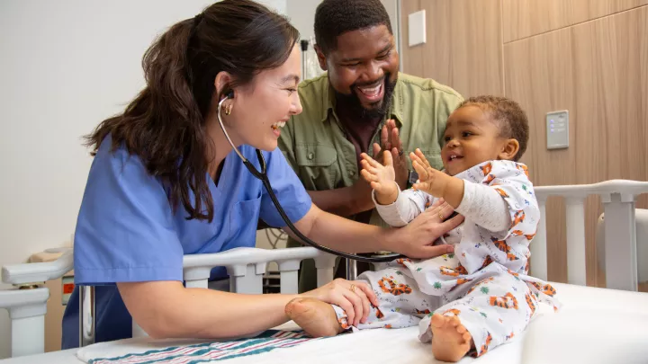 happy toddler held by dark skinned man in green shirt and examined by Asian female doctor