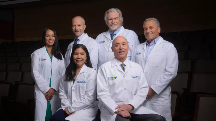 Seven researchers wearing white lab coats pose for a group picture in two rows against a dark background