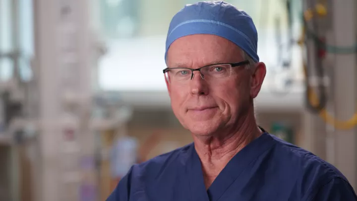 A man with light skin tone and glasses wears dark blue scrubs and a light blue surgical cap in front of a blurred background of medical equipment.