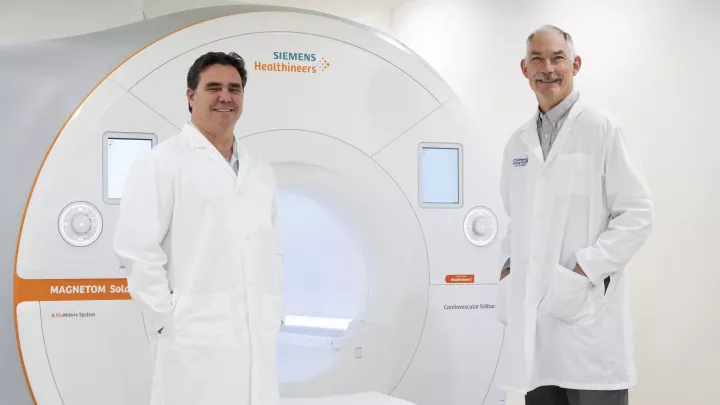 Two men wearing white lab coats stand on either side of a large white MRI machine. The machine has a large round tunnel-like opening.