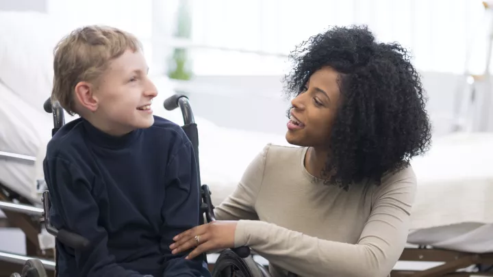 A light-haired boy in a wheelchair smiles at a dark-haired woman who is crouched down beside him in front of a white hospital bed.