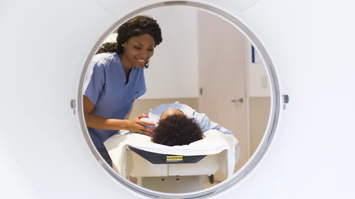 A nurse with medium skin tone and dark hair smiles at a child lying on a gurney who is about to enter an MRI machine. The scene is shown through the circular opening of the MRI.