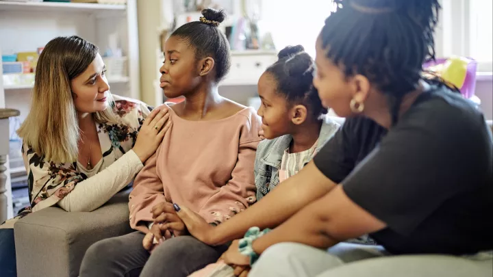 Three siblings sit on a couch while comforted by a friend. 