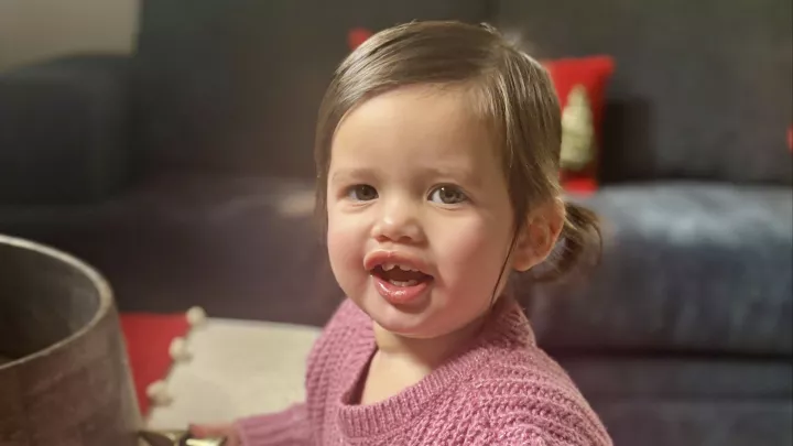 A toddler stands at a living room coffee table smiling at the camera