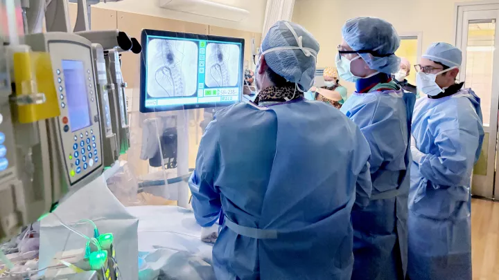 Three men in blue scrubs, caps and masks gather round a large computer monitor in a hospital room, with equipment in the foreground.