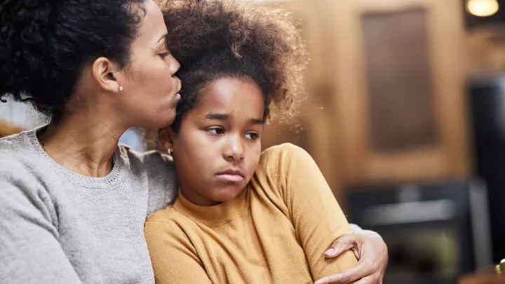 A dark skin-toned girl looks sad. A dark skin-toned woman hugs her and kisses her head.