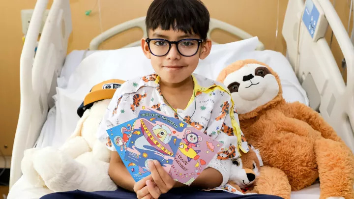 Young patient with medium skin tone, dark hair and glasses holds brightly colored Valentine's Day cards in their hand as they sit in a hospital bed