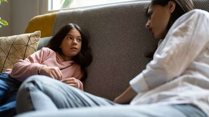 A school-aged girl and her mother have a serious conversation while reclining on the couch. 