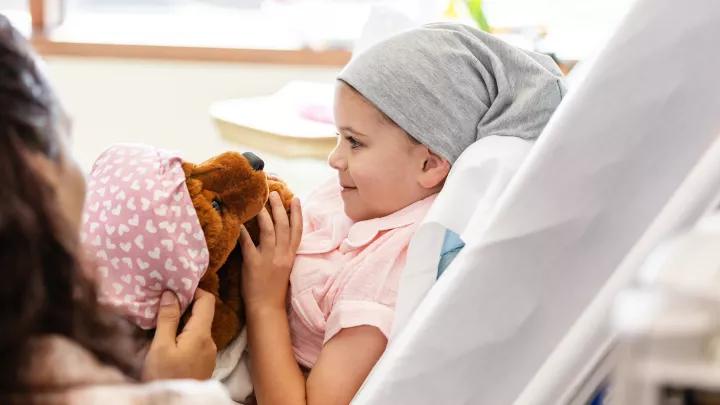 A child in a hospital bed wearing pajamas and a hat holds her stuffed dog wearing a hat. 