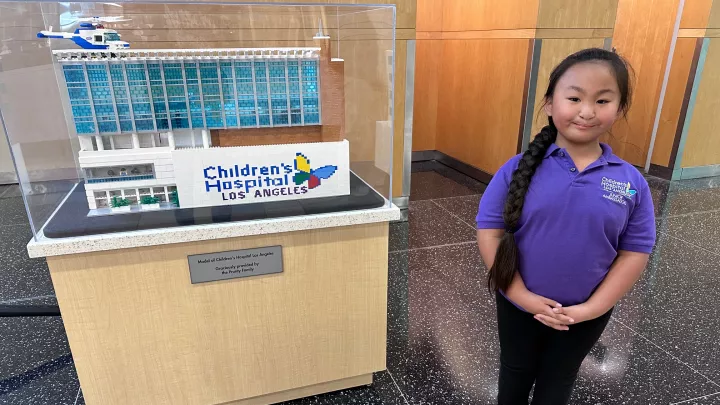 Young girl with long, dark, braided hair to the side wearing a purple polo shirt and standing next to a Lego model in a lobby.