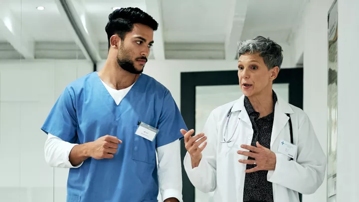 A medium-light skin toned woman with short gray hair wearing a lab coat and stethoscope talks to a medium skin toned man in blue scrubs as they walk. 