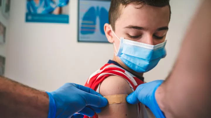Gloved hands apply a bandaid to the shoulder of a young boy wearing a blue medical procedure mask.
