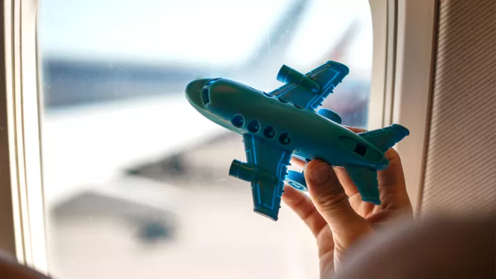 Child's hand holding a blue toy airplane next to the window of a passenger plane.