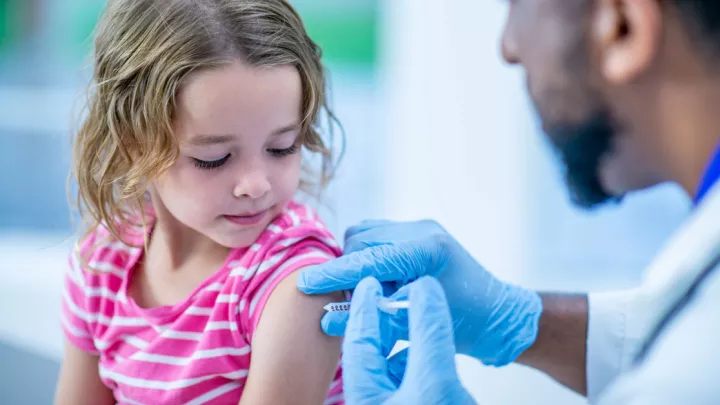 Young girl receives a vaccine shot in her left arm by a doctor wearing a lab coat and medical gloves.