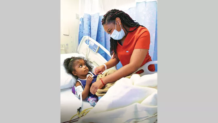 Nurse wearing a procedure mask looking at and helping a young patient who is in a hospital bed.