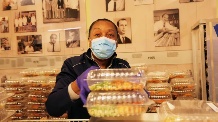 Masked woman with dark skin and wearing surgical mask prepares boxed meals