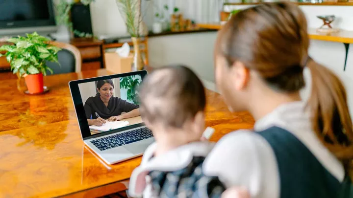 A woman holds a baby while on a video call with a doctor through a laptop.