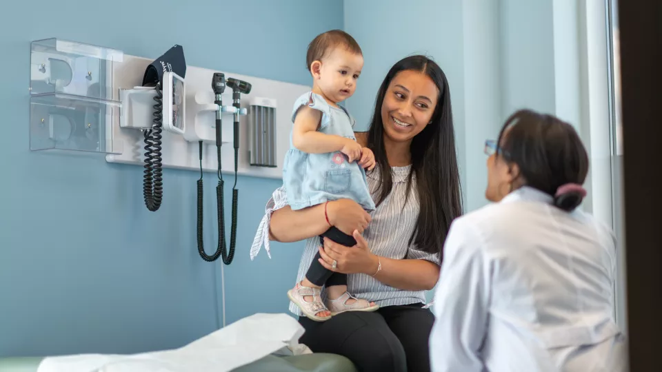 A young woman with medium skin tone sits on exam table holding a toddler. A doctor with medium-dark skin tone is speaking to them.