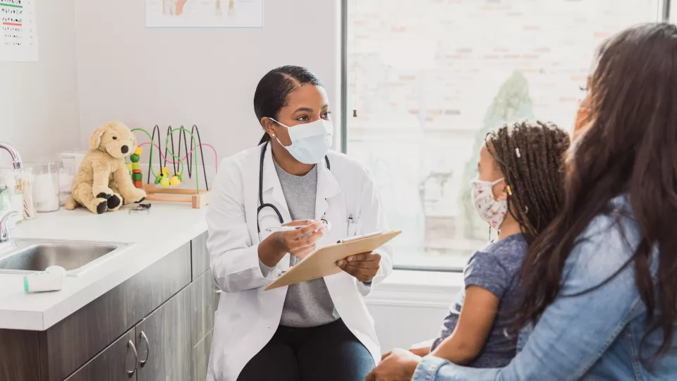 Female pediatrician with dark skin tone wearing surgical mask and white lab coat talks with patient's mother