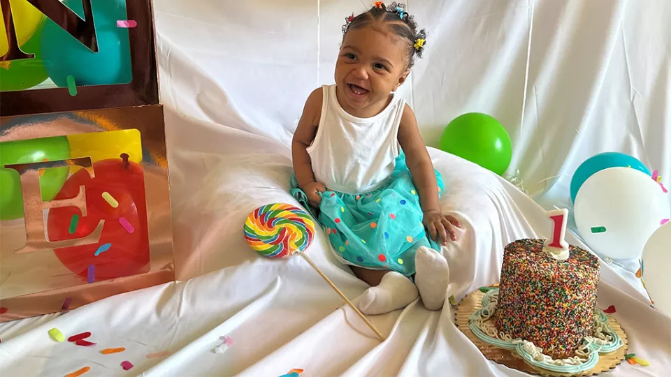 Ciara, a medium skin-toned toddler, smiles as she sits next to a rainbow swirl lollipop and a birthday cake topped with a number one candle