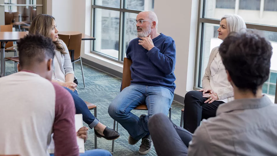 A group of adults sit on chairs in a circle, while one of them speaks to the rest of the group.