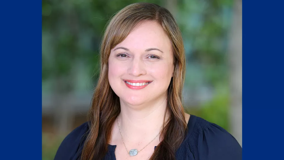 Headshot of smiling woman with light skin tone and brown hair wearing gemstone necklace and dark blue blouse against blurred outdoor background