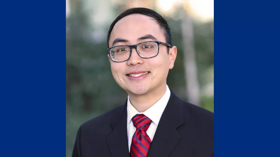 Headshot of a smiling man with light skin tone and short dark hair wearing glasses, dark suit jacket and red and blue striped tie against a blurred outdoor background